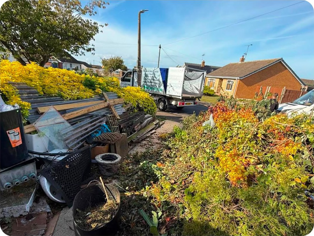 A front garden crowded with overgrown shrubs, old fence panels, pots and rubbish