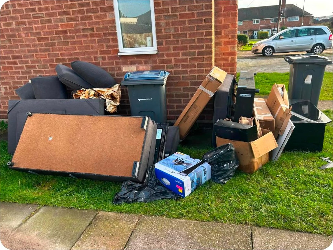An old sofa, bed base, boxes and rubbish bags piled on a front lawn beside the bins